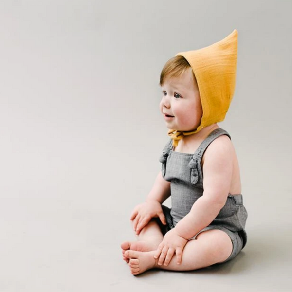 baby in grey overalls wearing mustard pixie bonnete 