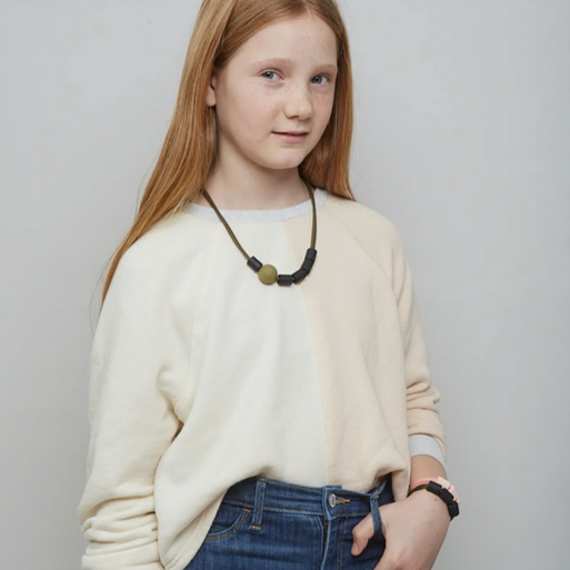 A child wearing a beige sweater and blue jeans, showcasing a black and green teething necklace.
